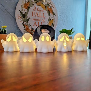 May include: Five white ceramic ghost figurines with glowing yellow eyes and mouths. The ghosts are arranged in a row on a wooden table.