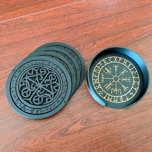 May include: A set of black coasters with intricate, carved designs. One coaster features a compass-like symbol in gold, while the others display a geometric pattern. The coasters are arranged on a wooden surface.