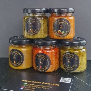 May include: A pyramid of five glass jars of gourmet sauces with black lids and labels. The labels read "Chez Fadi's" and include text like "Magic Sauce" and "Hot & Spicy Sauce." A business card with contact information is in front.