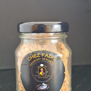 May include: A glass jar with a black lid filled with a brown spice blend. The label reads "Chez Fadis Feinkost Gewürze" with a small illustration of a woman and the text "Fish Mix".