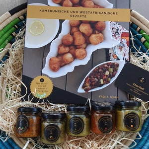 May include: A wicker basket filled with shredded paper and six small glass jars of hot sauce. The jars are labeled "Chez Fadi" and have different colored labels. The basket is sitting on a wooden table next to a cookbook titled "Kamerunische und Westafrikanische Rezepte" with a picture of fried dough balls on the cover.