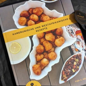 May include: A cookbook with a yellow cover featuring the text "Kamerunische und Westafrikanische Rezepte" and a white bowl filled with small, round, golden brown fried dough balls. The book also has a sticker that says "Mit mehr als 20 veganen Rezepten Vegan".