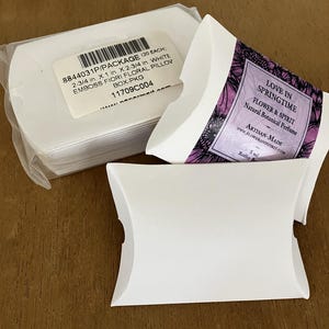 May include: A stack of white cardboard pillow boxes in a clear plastic sleeve, alongside two individual white pillow boxes. One box features a purple label with the text LOVE IN SPRINGTIME FLOWER & SPIRIT Natural Botanical Perfume.