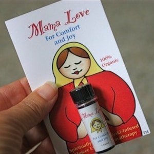 May include: A small glass bottle with a black cap and a label that reads "Mama Love For Comfort and Joy" is held next to a card with the same text and a cartoon illustration. The card also states "100% Organic". The bottle contains a purple liquid.
