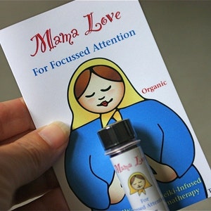 Könnte beinhalten: Eine weiße Karte mit dem Text "Mama Love" und "For Focussed Attention" in roter und blauer Schrift. Auf der Karte ist eine Comic-Illustration einer Frau mit braunen Haaren und einem gelben Kopftuch zu sehen. Vor der Karte steht eine kleine Glasflasche mit schwarzem Verschluss und einem Etikett mit der Aufschrift "Mama Love".