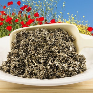 May include: A white scoop filled with dried, light green, and gray herbs on a white plate. The plate is on a light brown wooden surface. A background of red poppies and yellow flowers is blurred.