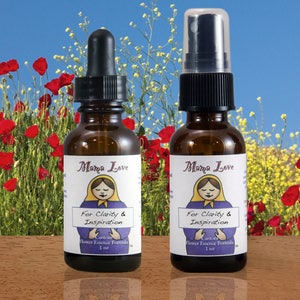 Op de afbeelding: Twee bruine glazen flessen met Mama Love bloemenessence. E&eacute;n heeft een druppelaar, de andere een sproeikop. Elke fles heeft een etiket met een Matroesjka-pop illustratie en de tekst "For Clarity & Inspiration". De achtergrond toont rode klaprozen en gele bloemen.