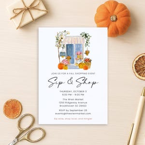 May include: A white invitation with the text "Sip & Shop" and event details. The invitation features a watercolor illustration of a shop with pumpkins and flowers. Other items include a gift box, scissors, a pen, and a dried orange slice.