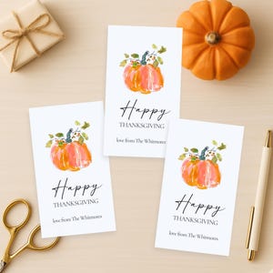 K&ouml;nnte beinhalten: Drei wei&szlig;e Thanksgiving-Karten mit Aquarellk&uuml;rbissen und dem Text "Happy Thanksgiving". Ein kleiner orangefarbener K&uuml;rbis und eine verpackte Geschenkbox sind ebenfalls vorhanden. Ein goldener Stift und eine Schere sind ebenfalls sichtbar.