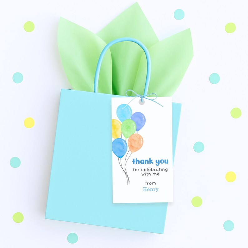 Puede incluir: Una bolsa de regalo azul claro con un asa a juego, llena de papel de seda verde. Una etiqueta blanca con globos de acuarela y el texto "thank you for celebrating with me from Henry" est&aacute; adjunta.