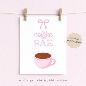May include: A white sign with the words "coffee BAR" in pink script and a pink bow illustration. A pink coffee cup with brown coffee is at the bottom. The sign is held by two wooden clips. The sign is 8x10 inches.