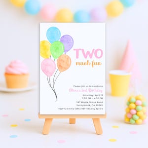 May include: A white invitation card on a small wooden easel, featuring watercolor balloons in various colors. The text reads "TWO much fun" and invites guests to Olivia's 2nd birthday party. Additional details include the date, time, and location.