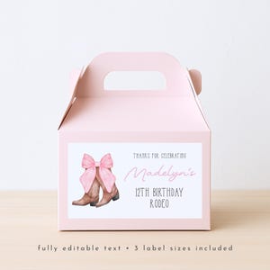 May include: A pale pink party favour box with a white label featuring a pink bow and brown cowboy boots. The label reads "Madelyn's 12th Birthday Rodeo" in pink script. The box has a handle and is on a light wood surface.