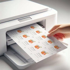 K&ouml;nnte beinhalten: Ein Bogen bedruckter Etiketten mit Aquarellk&uuml;rbissen und dem Text "Happy Thanksgiving". Die Etiketten werden von einem wei&szlig;en Drucker gedruckt. Die Etiketten werden von einer Hand gehalten.
