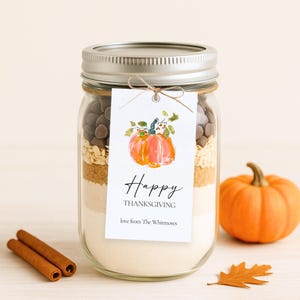 K&ouml;nnte beinhalten: Ein klares Glas mit Zutaten zum Backen von Keksen, darunter Schokoladenst&uuml;ckchen und Haferflocken. Ein Etikett mit einem Aquarellk&uuml;rbis und den Worten "Happy Thanksgiving" ist am Glas befestigt. Ein kleiner oranger K&uuml;rbis und Zimtstangen liegen daneben.