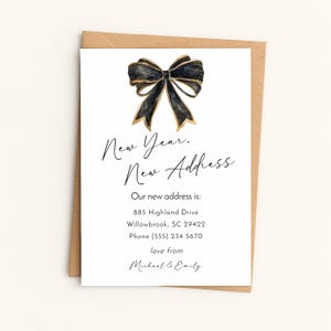 May include: A white card with a black and gold bow illustration. The card reads "New Year, New Address" and includes a new address, phone number, and names. The card is set against a brown envelope.