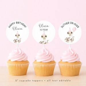 Puede incluir: Tres cupcakes con glaseado rosa y envoltorios amarillos. Cada uno tiene un adorno circular blanco con una ilustración de serpiente con un gorro de fiesta. El texto en los adornos dice "Happy Birthday Olivia", "Olivia is Four" y "Slither on Over".