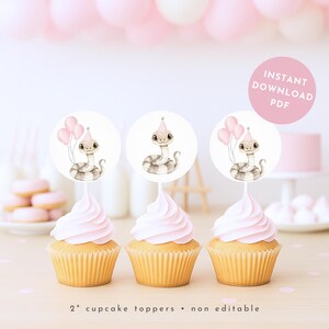 Pode incluir: Três cupcakes com cobertura rosa e toppers de cupcake com uma ilustração em aquarela de uma cobra usando um chapéu de festa. Os toppers têm 5 cm de diâmetro. O fundo inclui balões rosa e um bolo.