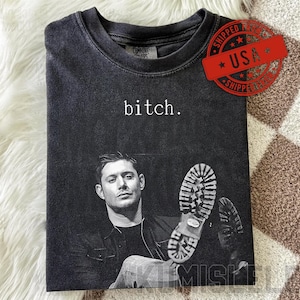 Könnte beinhalten: Dunkelgraues T-Shirt mit dem Wort "bitch" in Weiß. Das Shirt zeigt ein Schwarz-Weiß-Bild eines Mannes und Schuhsohlen. Ein roter "USA"-Stempel ist sichtbar.