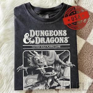 May include: Dark grey t-shirt with the text "DUNGEONS & DRAGONS" and a fantasy illustration of a dragon and warrior. The shirt is folded, with a vintage, distressed appearance.