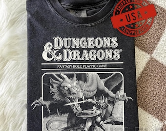 Może przedstawiać: Ciemnoszara koszulka z napisem "DUNGEONS & DRAGONS" i fantastyczną ilustracją smoka i wojownika. Koszulka jest złożona i ma wygląd vintage.