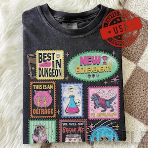 May include: Dark grey t-shirt with colourful graphic designs resembling postage stamps. The stamps feature phrases like "Best in Dungeon," "New Achievement," and "This is an Outrage." A red "USA" stamp is visible.
