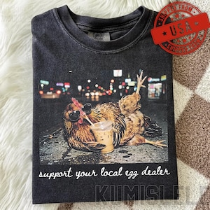 May include: A charcoal grey t-shirt featuring a graphic of a chicken wearing black sunglasses and holding a plastic cup with a straw. The text below the graphic reads, support your local egg dealer, in a white, cursive, lowercase font.