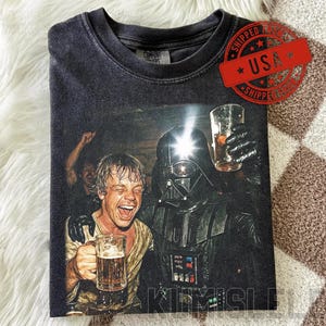 Könnte beinhalten: Dunkelgraues T-Shirt mit einem humorvollen Aufdruck von Darth Vader und einem Mann, der mit Bierkrügen anstößt. Das Bild hat einen Vintage- und Used-Look. Das T-Shirt hat einen Rundhalsausschnitt und kurze Ärmel.