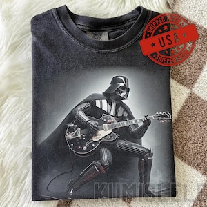 Op de afbeelding: Donkergrijs T-shirt met een zwart-wit afbeelding van Darth Vader die gitaar speelt. De afbeelding bevat een rode "USA" stempel. Het shirt is opgevouwen op een witte, vachtachtige ondergrond.