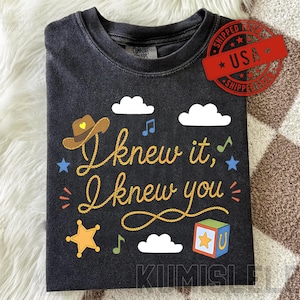 Puede incluir: Una camiseta gris carbón de manga corta con el texto "I knew it, I knew you" en una fuente amarilla estilo cuerda. El diseño incluye un sombrero de vaquero marrón, nubes blancas, notas musicales azules y verdes, una estrella de sheriff amarilla.