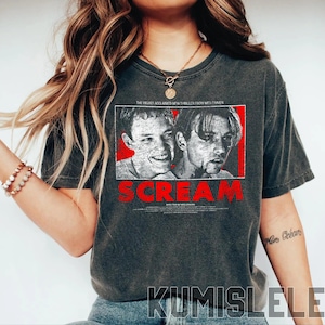 May include: Dark gray t-shirt featuring a graphic from the movie "Scream." The design includes a red rectangle with two black and white portraits and the title "SCREAM" in red. Text above the image reads "THE HIGHLY ACCLAIMED NEW THRILLER FROM WES CRAVEN."