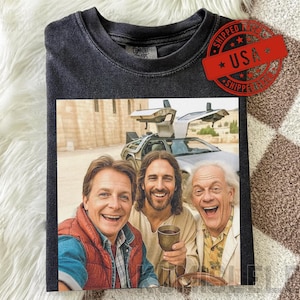 May include: Dark gray t-shirt featuring a colorful image of three smiling men, one holding a chalice, with a DeLorean car in the background. The shirt has a distressed look and a red "USA" stamp.