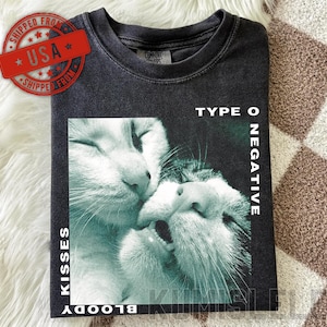 以下が含まれることがあります： チャコールグレーのクルーネックTシャツ。寄り添う2匹の猫のグラフィックがプリントされています。右側に「TYPE O NEGATIVE」、左側に「KISSES」という文字が、それぞれ白いサンセリフ体のフォントで縦方向にプリントされています。