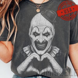 May include: A charcoal grey t-shirt featuring a detailed black and white graphic print of a clown wearing a bowler hat and a ruffled collar. The clown is depicted with a wide, toothy grin and hands positioned to form a heart shape in front of the chest.