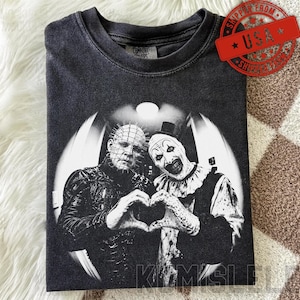 May include: A charcoal grey crew neck t-shirt featuring a black and white graphic print of Pinhead and Art the Clown forming a heart shape with their hands. The shirt has a vintage-style wash and a Comfort Colors brand label at the inner neckline.