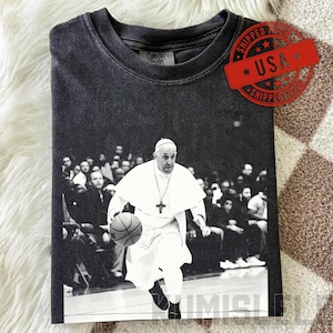 Op de afbeelding: Donkergrijs t-shirt met een zwart-wit afbeelding van de paus die basketbal speelt. De afbeelding is gedrukt op een voorgewassen t-shirt. Het shirt heeft een ronde hals en korte mouwen. Een rode stempel met "SHIPPED USA" is zichtbaar.