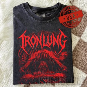 Puede incluir: Camiseta negra doblada con un diseño gráfico rojo. El texto "IRONLUNG" en fuente gótica está sobre la boca de una criatura. La camiseta tiene un aspecto vintage y desgastado.