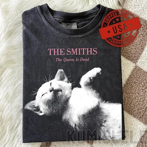 Puede incluir: Camiseta gris oscuro con el texto rosa "THE SMITHS" y "The Queen Is Dead". La camiseta tiene una imagen en blanco y negro de un gato. Una pegatina roja "SHIPPED USA" está en la parte superior derecha.