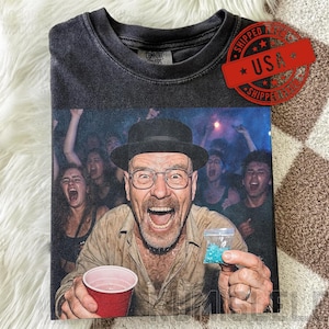 May include: Dark gray t-shirt featuring a vibrant image of a man in a hat and glasses, holding a red cup and a bag of blue crystals, with a crowd in the background. The shirt has a distressed look.