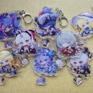 May include: A collection of anime-style character keychains. Each keychain features a unique character design with vibrant colors, including blue, pink, and yellow. The keychains are attached to silver-colored metal rings.