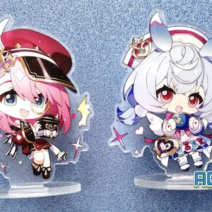 May include: Two anime-style character acrylic stands. The stand on the left features a pink-haired character with a red and black hat, holding a small object. The stand on the right features a white-haired character with blue hair accents, wearing a white dress with a blue bow and holding a heart-shaped object. Both stands have a clear base and feature glitter accents.