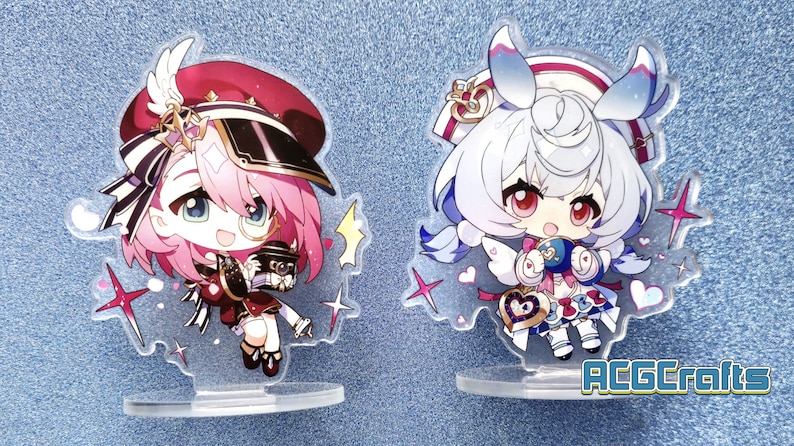 May include: Two anime-style character acrylic stands. The stand on the left features a pink-haired character with a red and black hat, holding a small object. The stand on the right features a white-haired character with blue hair accents, wearing a white dress with a blue bow and holding a heart-shaped object. Both stands have a clear base and feature glitter accents.