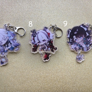 May include: Three anime-style acrylic keychains featuring chibi characters with silver key rings. The characters have colorful hair and outfits. Each keychain has a small charm attached. The background is a light tan color.