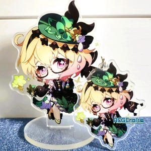 May include: Two acrylic charms featuring a cartoon character with blonde hair, wearing a green hat and a black and green dress. The character has pink cheeks and is holding a black object. The charms are on a clear acrylic stand. The larger charm is on a stand and the smaller charm is hanging from a chain.