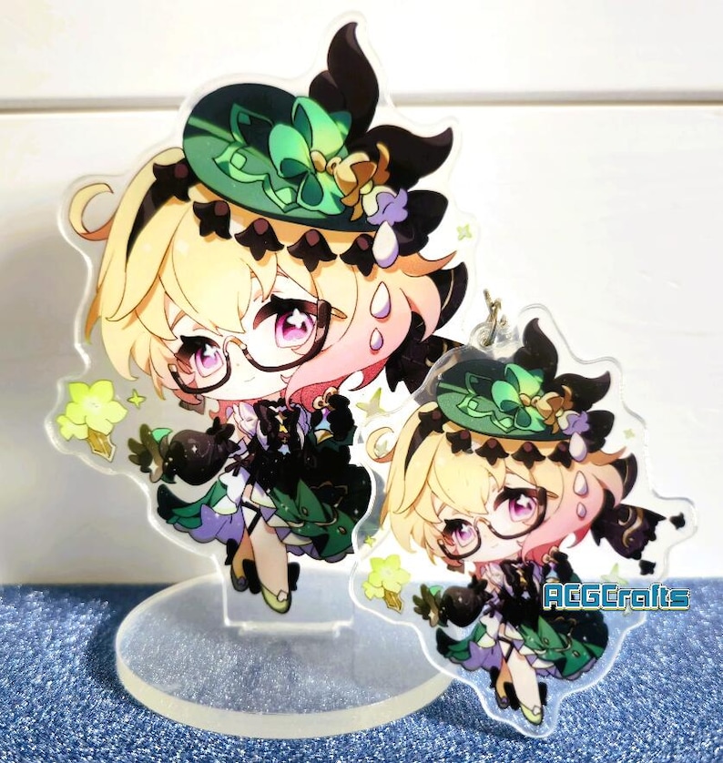 May include: Two acrylic charms featuring a cartoon character with blonde hair, wearing a green hat and a black and green dress. The character has pink cheeks and is holding a black object. The charms are on a clear acrylic stand. The larger charm is on a stand and the smaller charm is hanging from a chain.