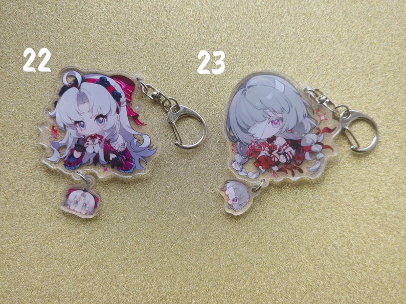 May include: Two anime-style acrylic keychains featuring female characters with large eyes and detailed outfits. One keychain has a white-haired character with pink accents, the other has a gray-haired character with red accents. Both have silver key rings.