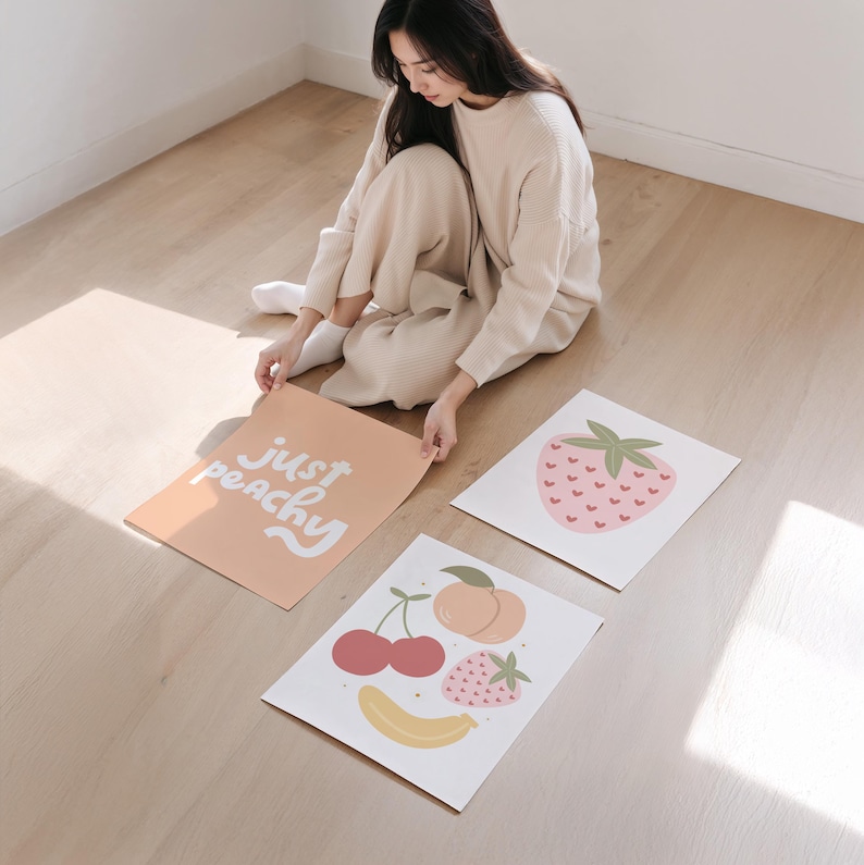 以下が含まれることがあります： フルーツのイラストが描かれた3枚の長方形のアートプリント。ピーチカラーのプリントには白い文字で「just peachy」と書かれています。白いプリントの1枚にはハート型の種を持つピンクのイチゴが描かれています。もう1枚の白いプリントには、桃、サクランボ、イチゴ、黄色いバナナが描かれています。