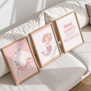 以下が含まれることがあります： 明るい木製フレームに入った3枚の壁掛けアートプリント。1枚目はピンクの背景に真珠と白い花が描かれた白い貝殻、2枚目は花を持つピンクのマーメイド、3枚目には「MAKE WAVES LITTLE MERMAID」という文字がデザインされています。