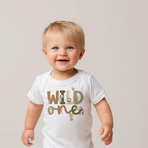 May include: A white baby bodysuit featuring the text "WILD ONE" in brown, green, and tan. The letters are adorned with animal illustrations, such as a giraffe, lion, and monkey. The baby is smiling.