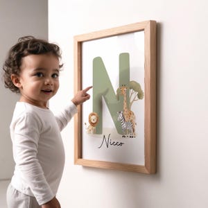 May include: A framed print with a light wood frame, featuring a green letter 'N' and safari animals: a lion, giraffe, and zebra. The name 'Nicco' is written in script below the letter. A child points at the print.